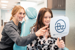 Girl smiling with orthodontic assistant, holding BZ Orthodontics sign, in modern dental office, showcasing positive patient experience at Lansdale orthodontist.