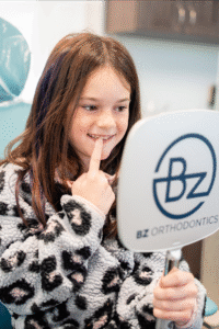 Girl smiling and pointing at her teeth while looking in a mirror with the BZ Orthodontics logo, showcasing a positive orthodontic experience related to braces and Invisalign in Lansdale, PA.