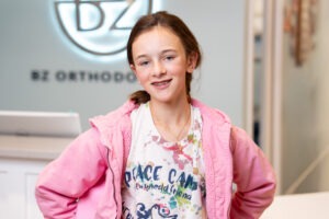 Girl smiling with orthodontic assistant, holding BZ Orthodontics sign, in modern dental office, showcasing positive patient experience at Lansdale orthodontist.
