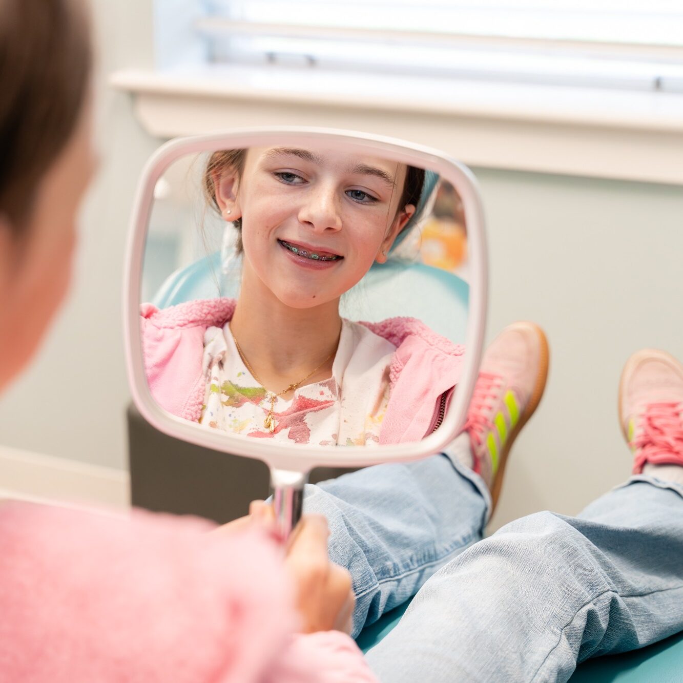 Girl with metal braces smiling in a dental chair, reflecting in a hand mirror, showcasing orthodontic treatment at BZ Orthodontics in Lansdale.